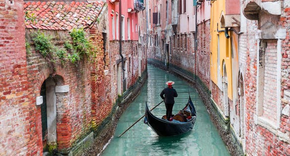 Venetian gondolier punting gondola through green canal waters of Venice, Italy.