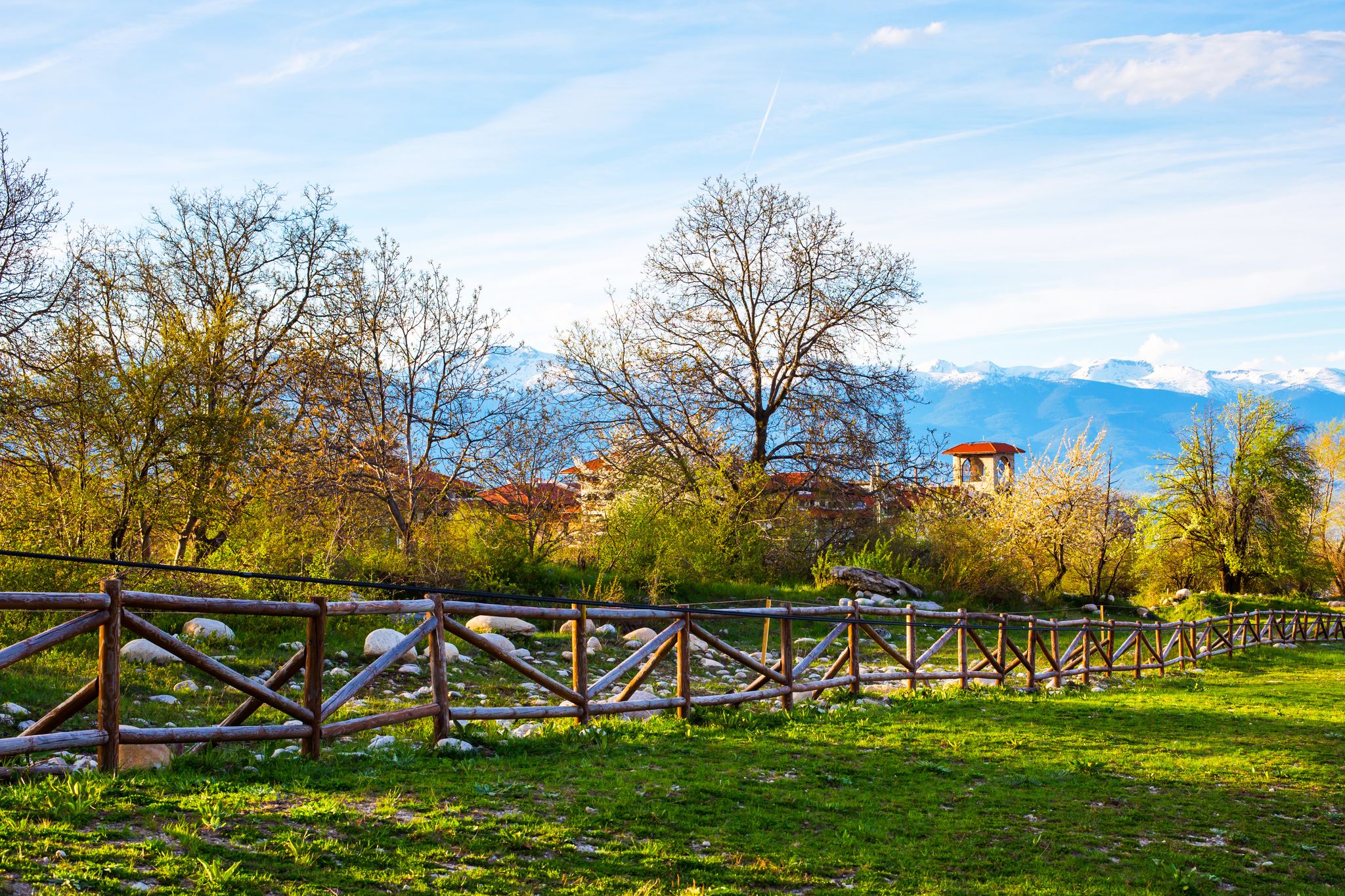 Photo of Landscape with the wooden fence, spring trees, part of the wooden tower of chalet and snowy peaks of mountains in Bansko, Bulgaria.