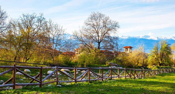 Photo of Landscape with the wooden fence, spring trees, part of the wooden tower of chalet and snowy peaks of mountains in Bansko, Bulgaria.