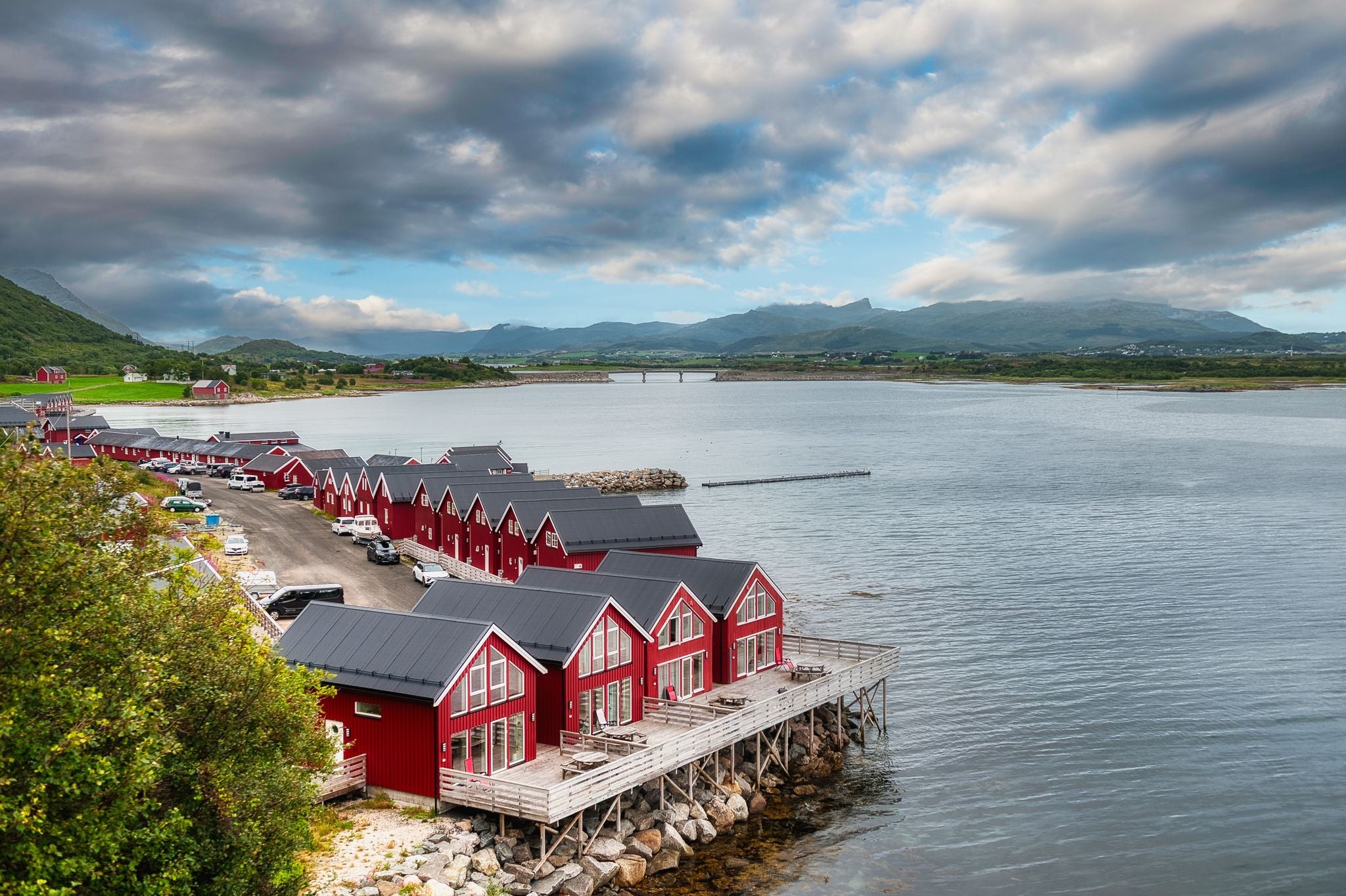 Rows of red seaside cabins on stilts sit along the calm harbor in Ballstad, near Leknes in Lofoten, Norway, with a scenic mountain backdrop under dramatic clouds.