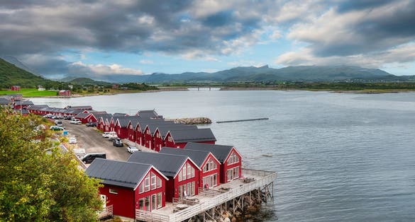 Rows of red seaside cabins on stilts sit along the calm harbor in Ballstad, near Leknes in Lofoten, Norway, with a scenic mountain backdrop under dramatic clouds.