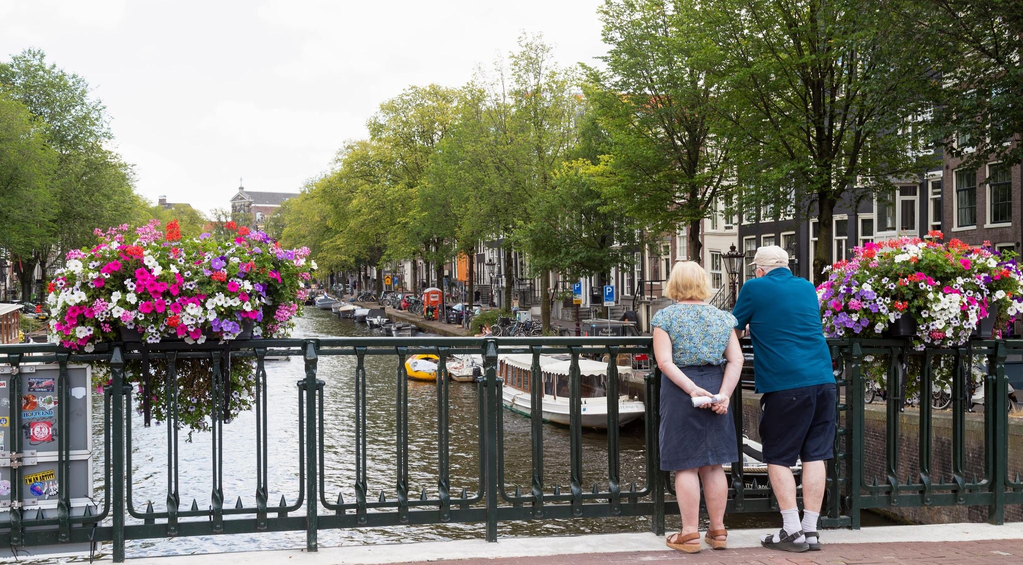 Older couple standing on a canal bridge in Amsterdam with flower baskets and boats below..jpg