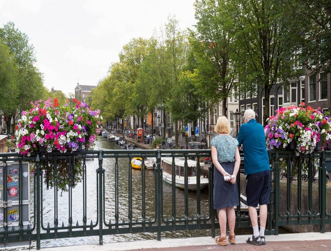 Older couple standing on a canal bridge in Amsterdam with flower baskets and boats below..jpg