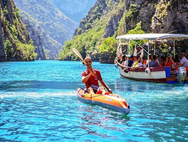 A man kayaks on the bright blue waters of Matka Canyon in North Macedonia, with a boat full of tourists nearby..jpg