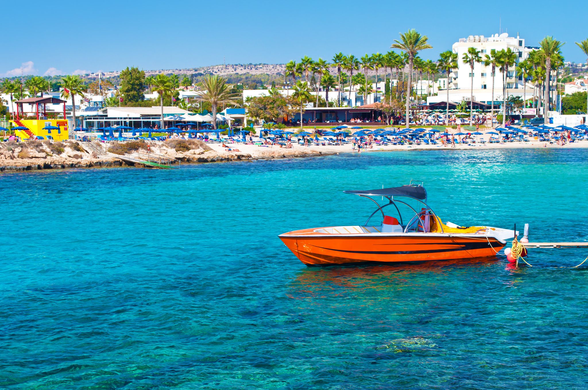 Photo of red speedboat near Vathia Gonia beach near Agia Napa, Cyprus. Sea with turquoise blue water, many green palm trees, houses on the background.