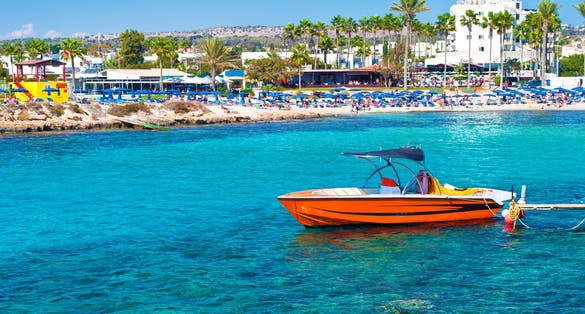 Photo of red speedboat near Vathia Gonia beach near Agia Napa, Cyprus. Sea with turquoise blue water, many green palm trees, houses on the background.