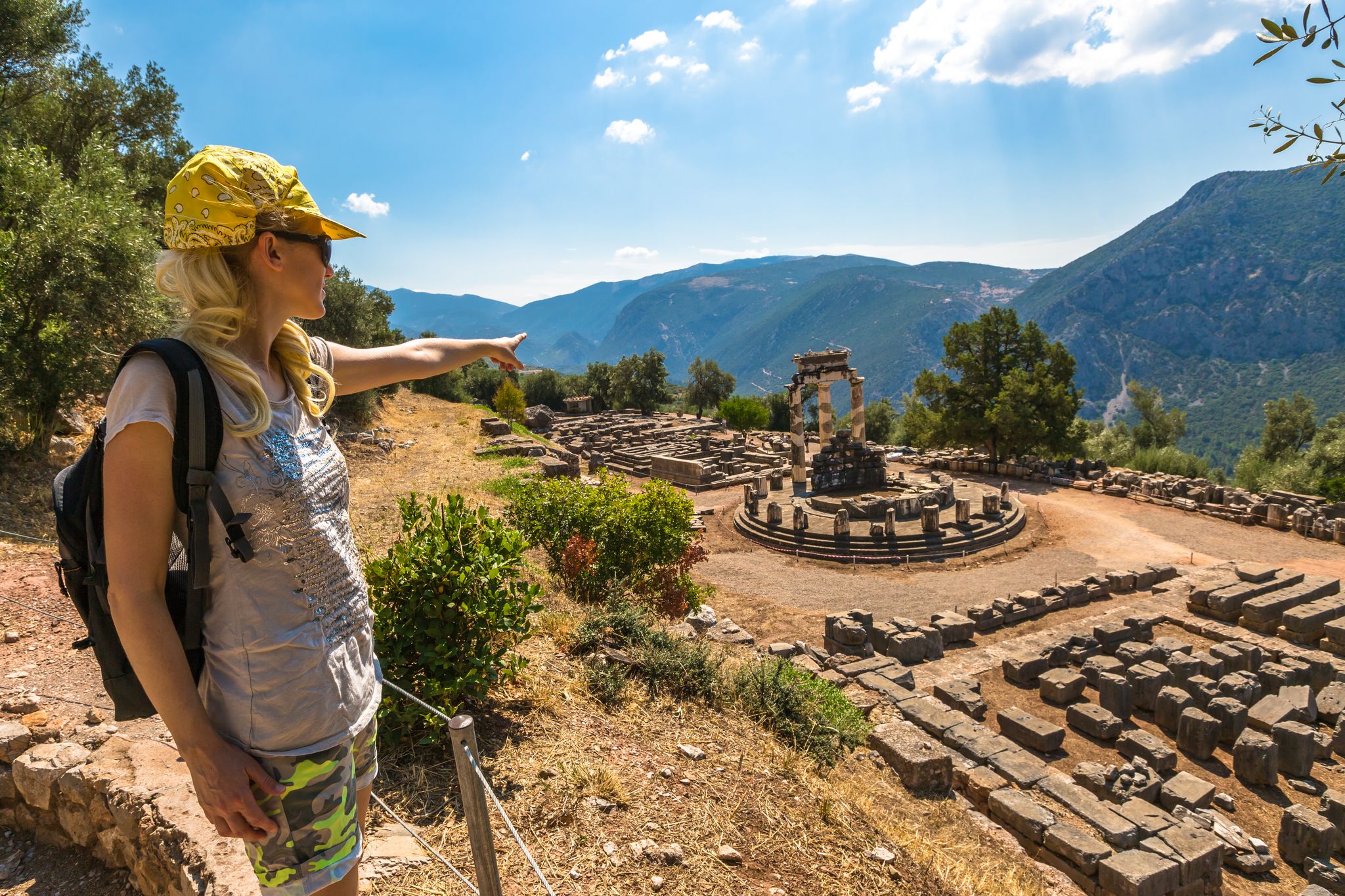 photo of A young tourist dressed in sportswear pointing to the spectacular ruins of the Tholos at the sanctuary of Athena Pronaia seen from an overlook, Delphi, Archaeological Site, Central Greece.,Delphi  Greece.