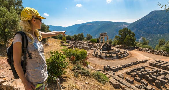 photo of A young tourist dressed in sportswear pointing to the spectacular ruins of the Tholos at the sanctuary of Athena Pronaia seen from an overlook, Delphi, Archaeological Site, Central Greece.,Delphi  Greece.