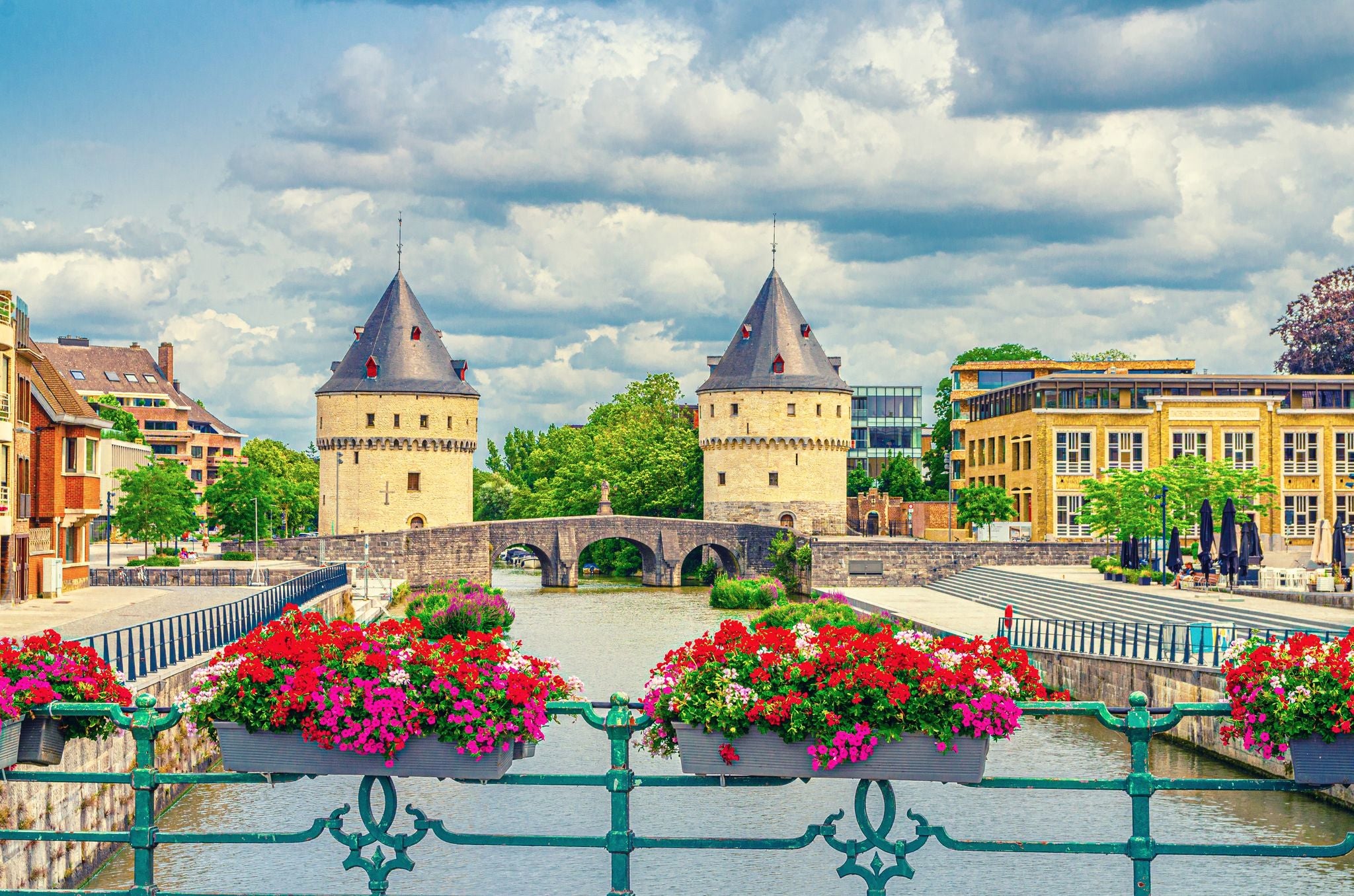 photo of view of Kortrijk cityscape with Lys river, Broel Towers Gothic style buildings and Bridge in historical centre, red flowers on bridge fence, Fortification towers, belgium landmark, Flemish Region, Belgium