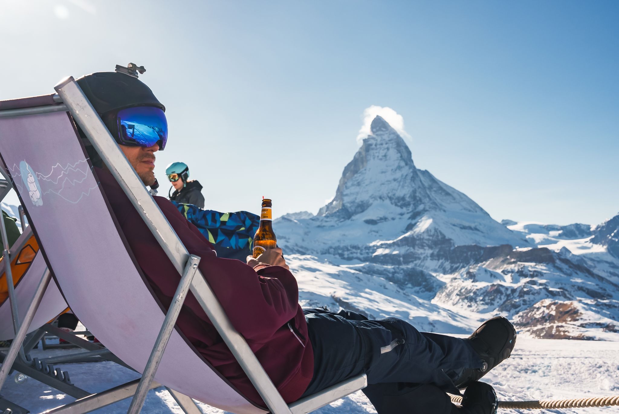photo of young snowboarder cheering with a beer after skiing day in a bar or a cafe at the Zermatt ski resort in Swiss Alps with a fabulous view of the Matterhorn.