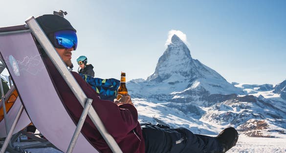 photo of young snowboarder cheering with a beer after skiing day in a bar or a cafe at the Zermatt ski resort in Swiss Alps with a fabulous view of the Matterhorn.