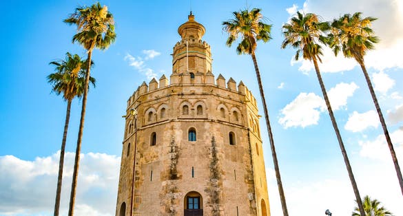 Photo of Torre del oro, historical limestone tower of gold under blue sky and palm tree in Seville, Spain.