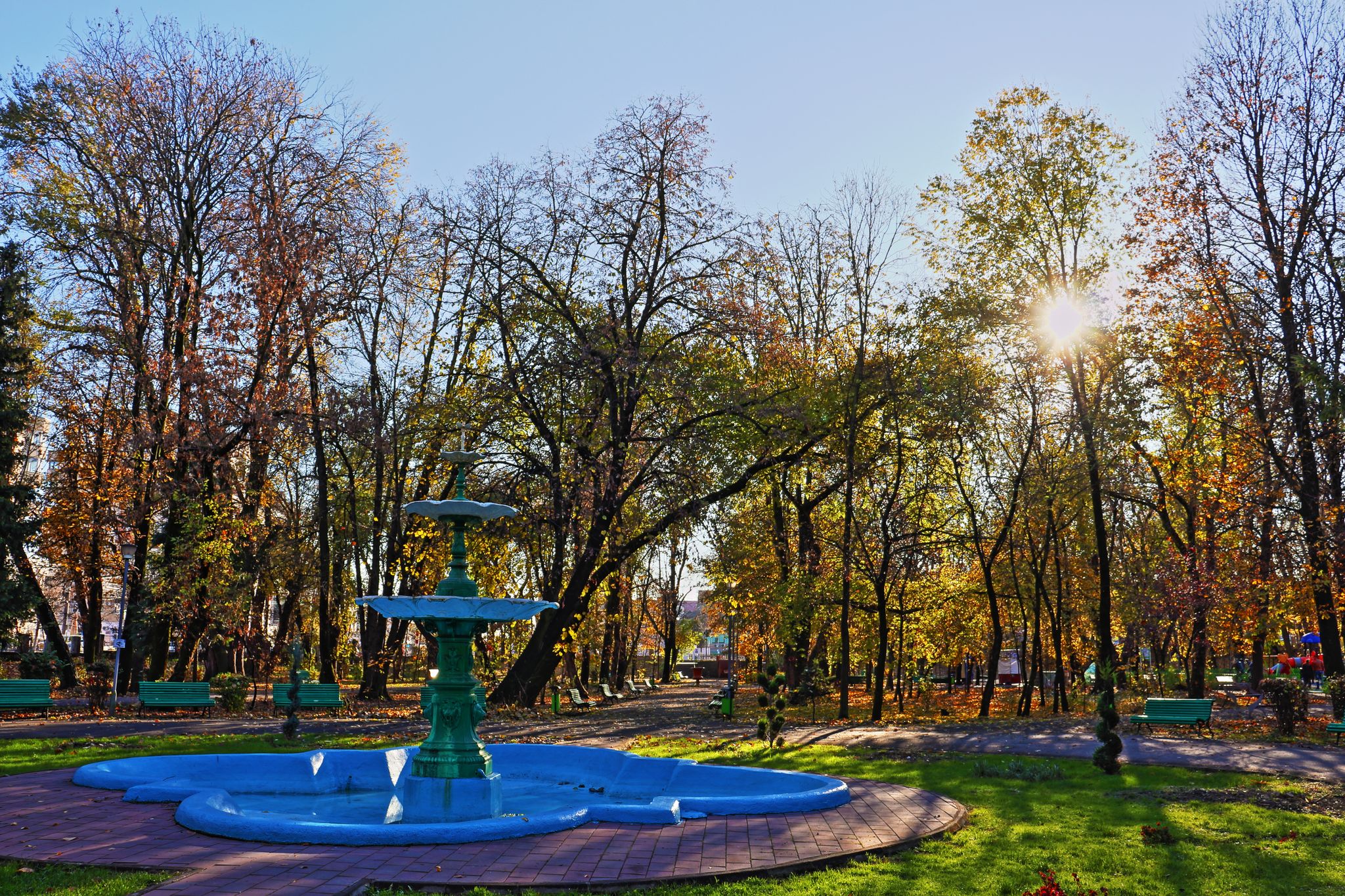 Beautiful park fountain and alley with benches in autumn pastel colors. High dynamic range photography. Sunny day outdoors. Sunrays through the tree crowns glowing bright.Targu Jiu tourist attraction