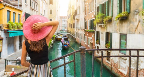 Elegant woman with red sunhat enjoys the view to a canal with passing by gondola in Venice, Italy.