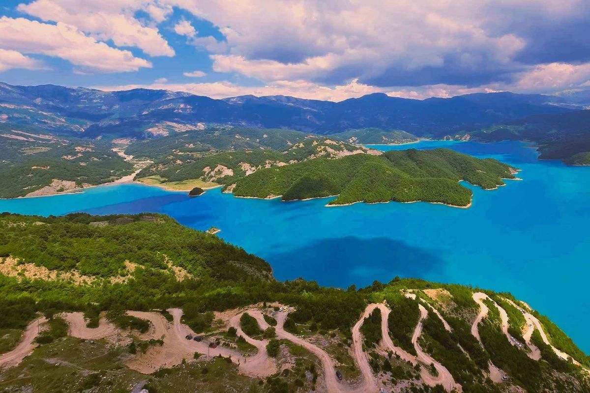 A curvy dirt road leads to Bovilla Lake, a blue lake surrounded by green hills and tall mountains..jpg