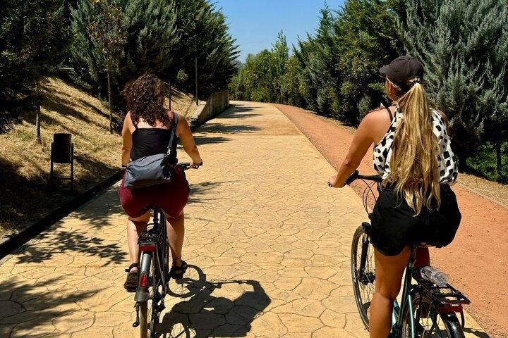Two women ride bicycles on a sunny path surrounded by trees during a bike tour in Tirana, Albania..jpg