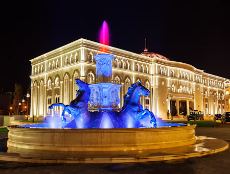 A beautiful fountain with horse statues glows in blue light at night in front of an elegant building in Skopje..png