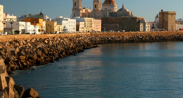 photo of the promenade in the city of Cadiz (Andalusia region, southern Spain) with the blue waters of the Atlantic Ocean and the Catedral de Cadiz building in the background.