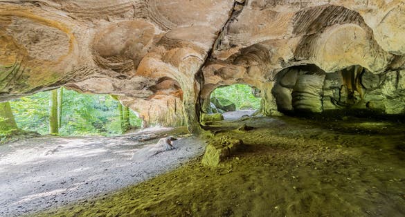 photo of rock formation huel lee or hohllay, hollow stone, was a quarry, circular patterns on the cave ceiling, uneven terrain and entry of sunlight, green trees in the background, Mullerthal trail, Luxembourg.