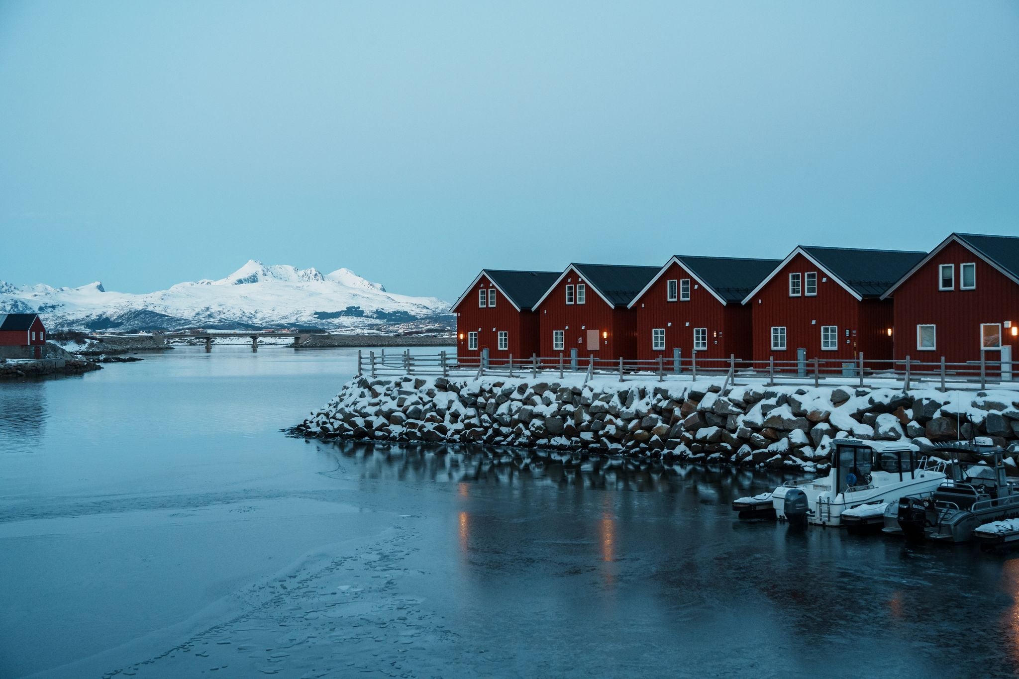 Red wooden cabins on stilts line the snowy waterfront in Ballstad, near Leknes in Lofoten, Norway, with frozen water and snow-covered mountains in the distance.