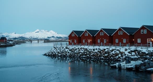 Red wooden cabins on stilts line the snowy waterfront in Ballstad, near Leknes in Lofoten, Norway, with frozen water and snow-covered mountains in the distance.