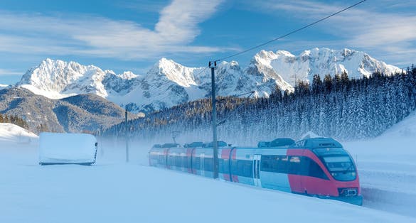 Photo of Winter scene of a train passing by a wooden barn in a valley covered by heavy snow with forests on the hillside & Karwendel mountains under blue sunny sky in background in Mittenwald, Bavaria, Germany.