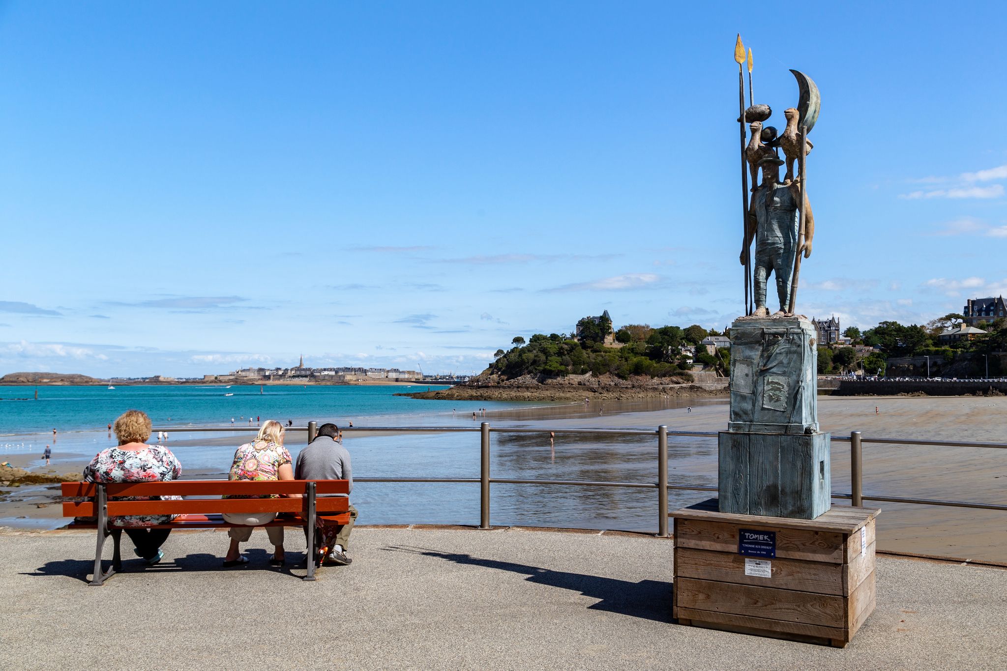 People rest on the beach promenade at the Man with Birds monument (by Tomek) at low tide.