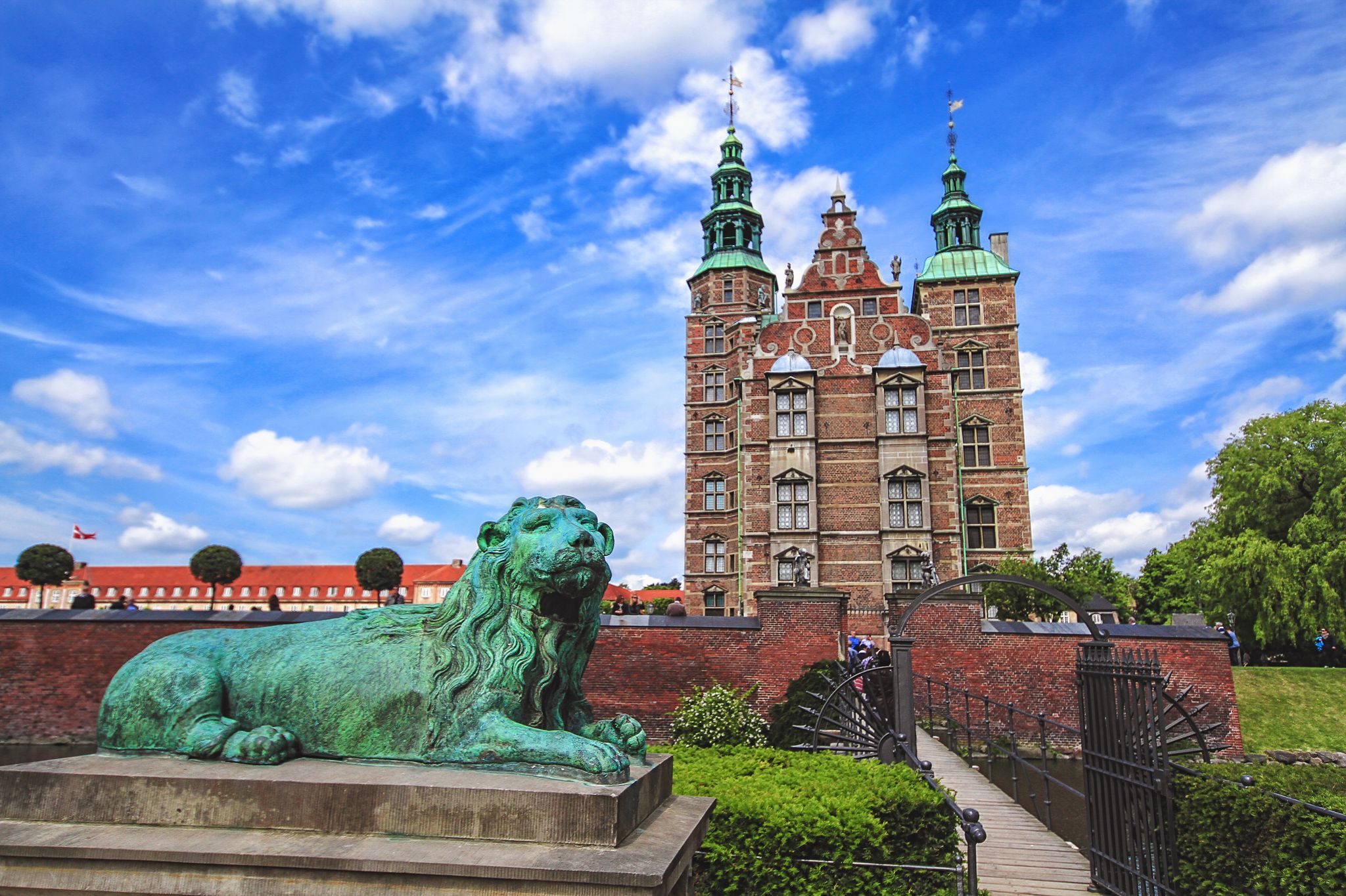 Photo of sculpture of lion against Rosenborg castle and blue sky. Statue is situated in front of bridge. 
