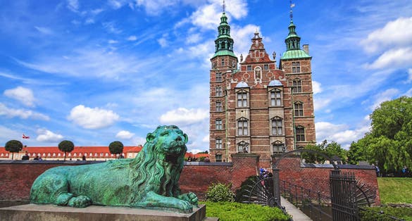 Photo of sculpture of lion against Rosenborg castle and blue sky. Statue is situated in front of bridge. 