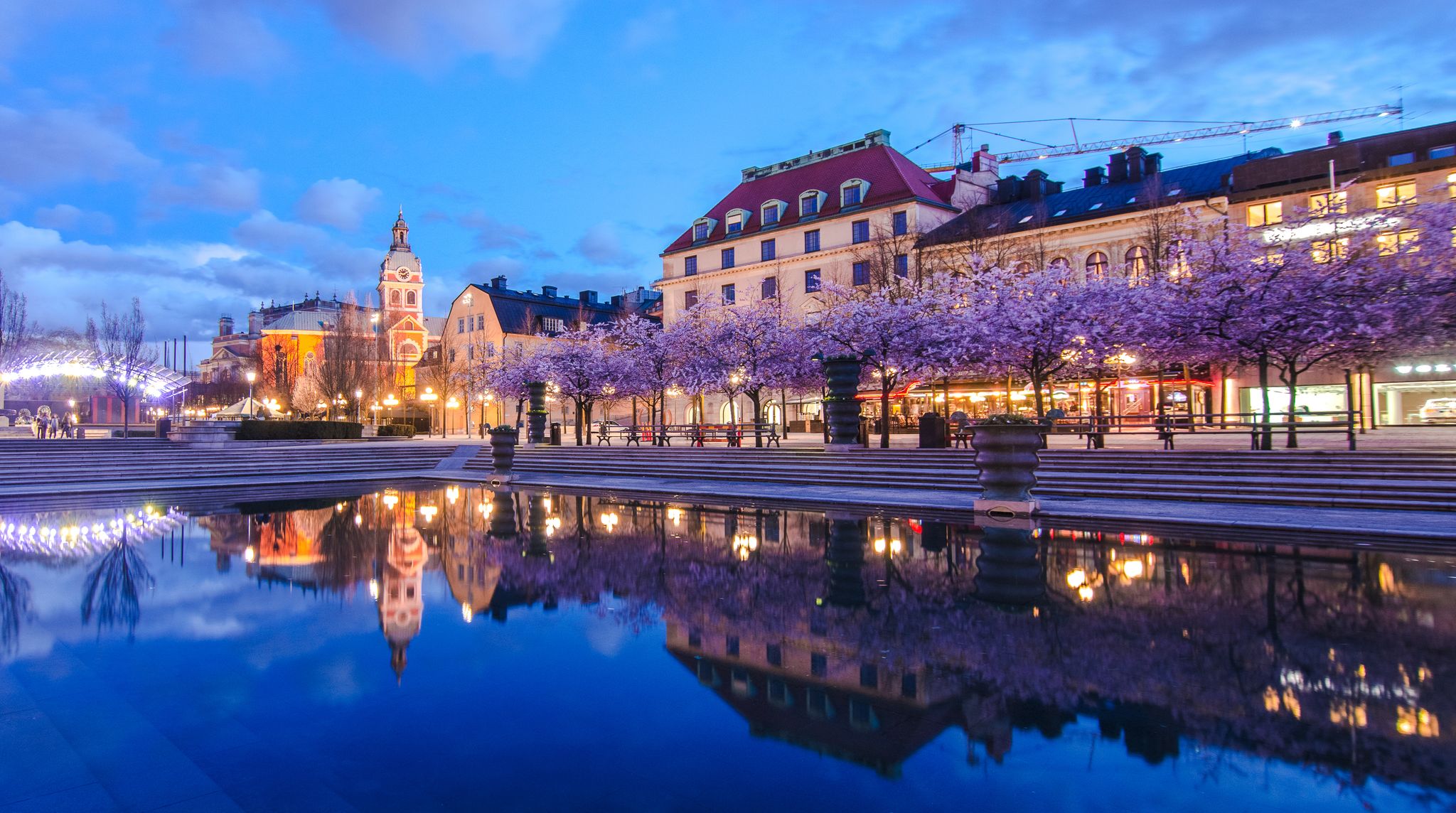 Photo of the famous meeting place Kungstradgarden and blooming cherry trees reflected in the water in Stockholm city center at dusk with historical buildings.