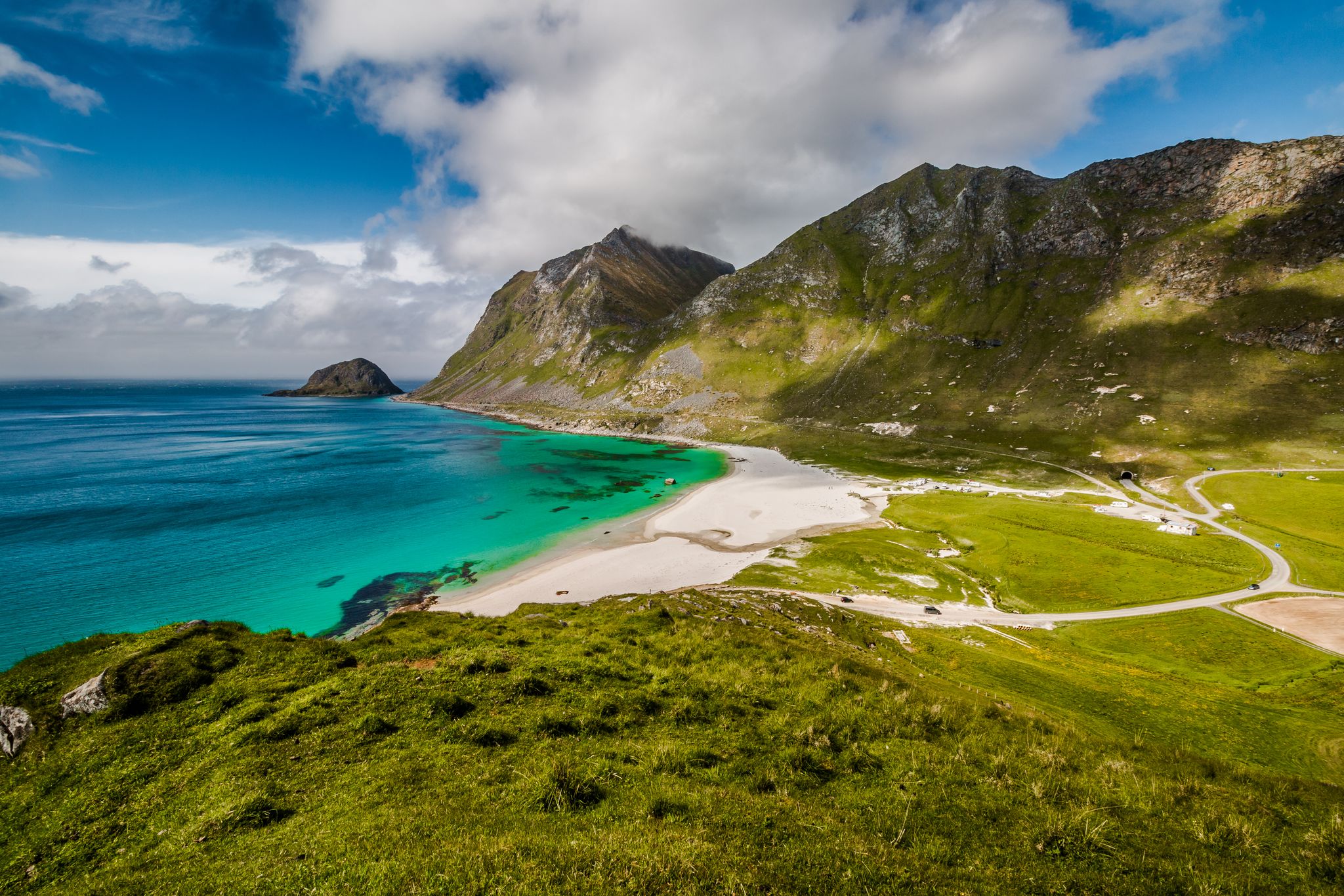 A view of Haukland Beach near Leknes in Lofoten, Norway, showing turquoise waters, white sand, and steep green mountains under a partly cloudy sky.