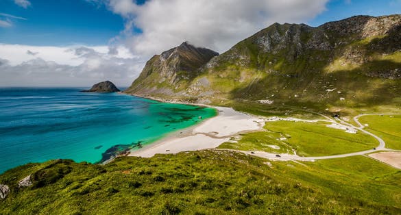 A view of Haukland Beach near Leknes in Lofoten, Norway, showing turquoise waters, white sand, and steep green mountains under a partly cloudy sky.