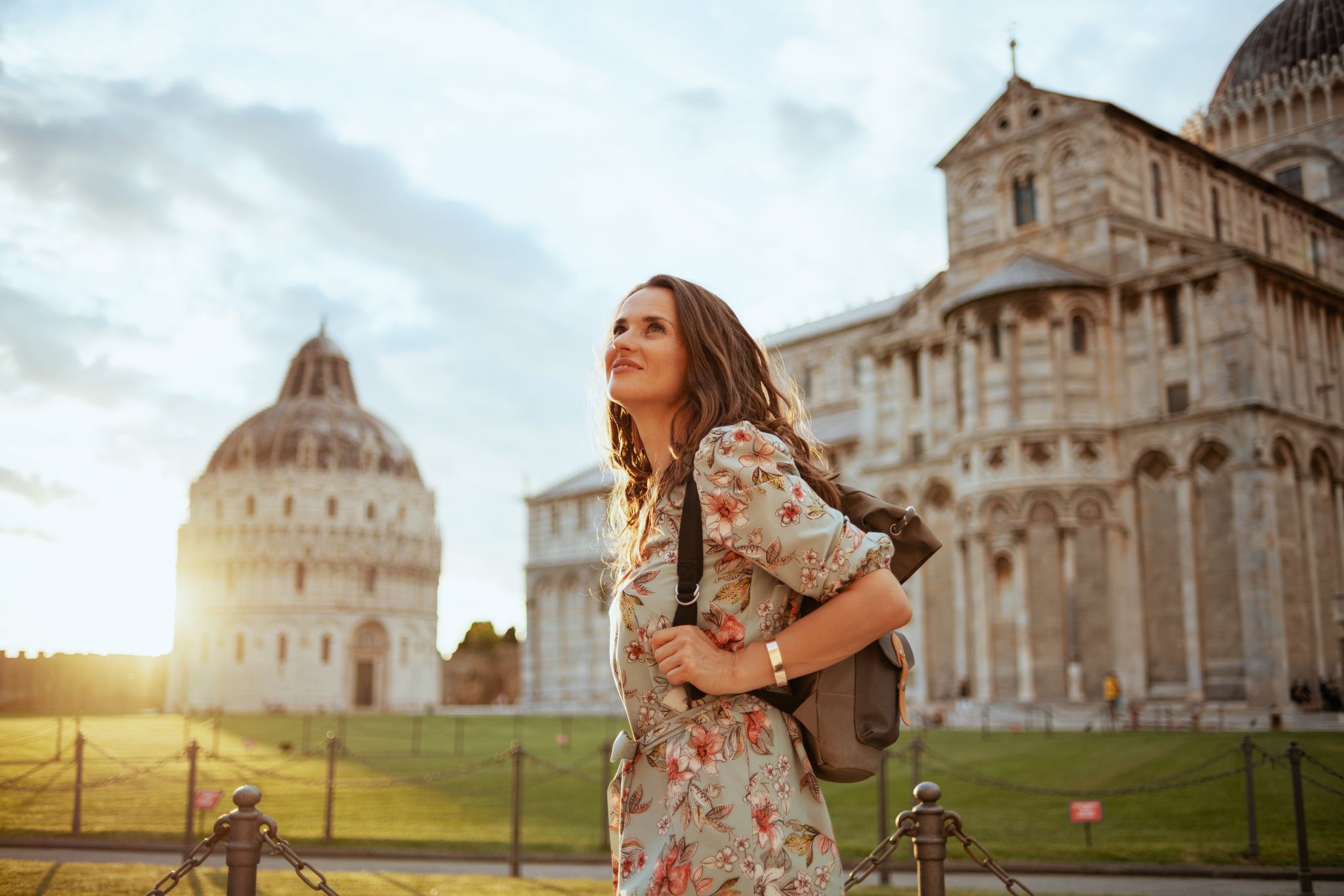 photo of smiling trendy traveler woman in floral dress with backpack enjoying promenade in piazza dei miracoli in Pisa, Italy.