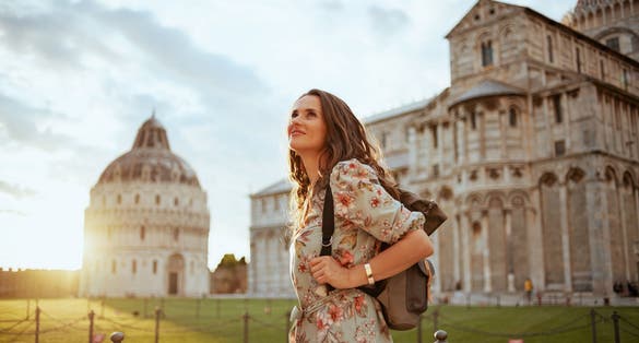 photo of smiling trendy traveler woman in floral dress with backpack enjoying promenade in piazza dei miracoli in Pisa, Italy.