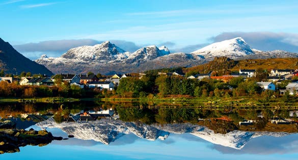 Snow-covered mountains reflect in the still fjord waters near Leknes, Norway, with a small village of colorful houses nestled at the base.