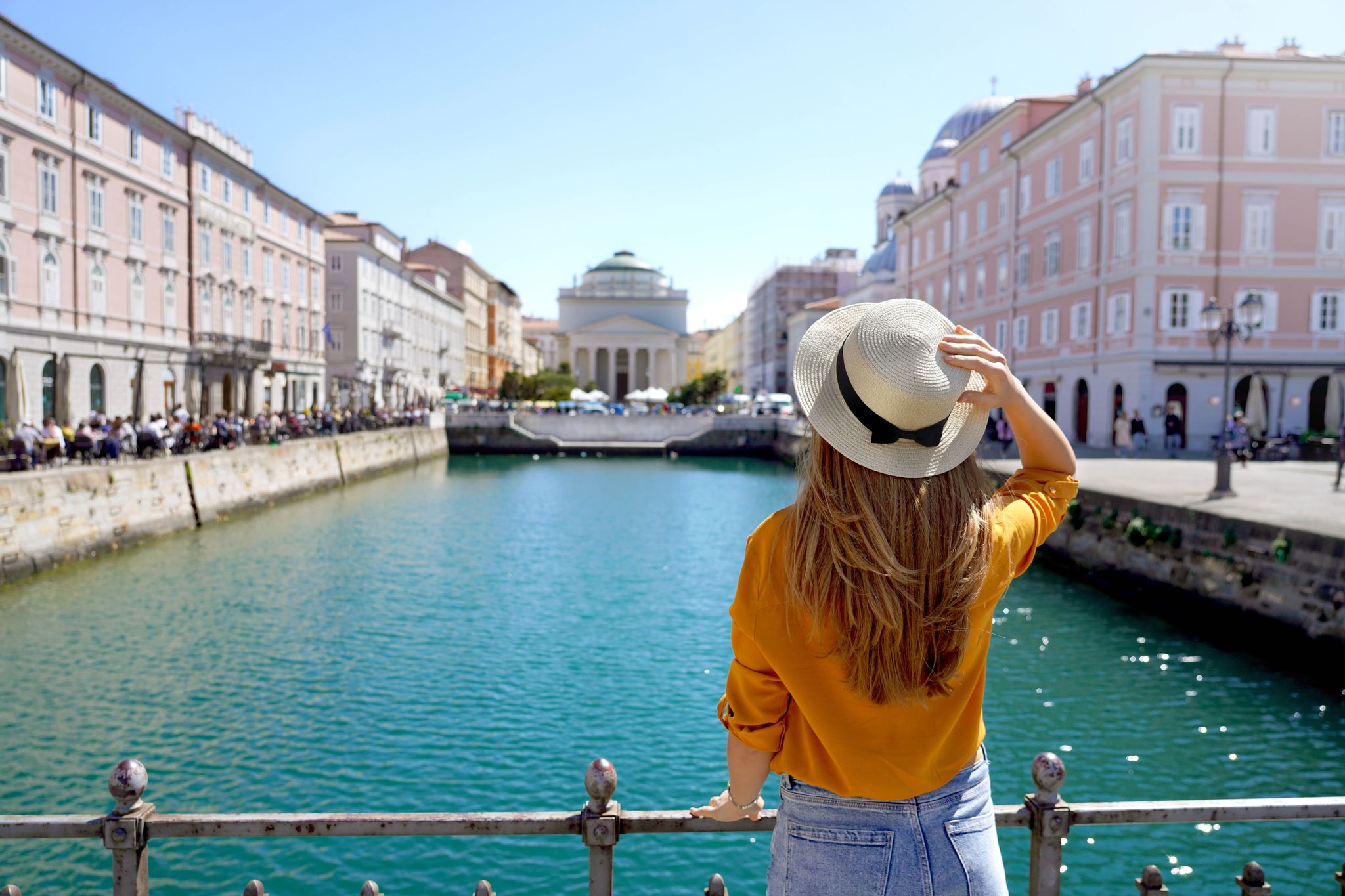 photo of view of Travel in Trieste, Italy. Back view of pretty girl holding hat looking at Sant Antonio Taumaturgo church on Grand Canal in Trieste, Italy.
