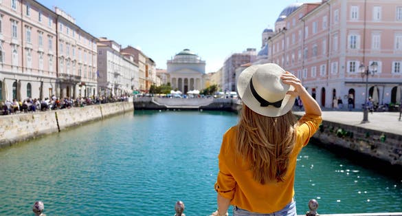 photo of view of Travel in Trieste, Italy. Back view of pretty girl holding hat looking at Sant Antonio Taumaturgo church on Grand Canal in Trieste, Italy.