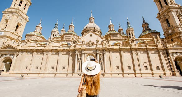 Photo of  tourist in sunhat standing back in front of the famous cathedral on the central square during the sunny weather in Zaragoza city, Spain.