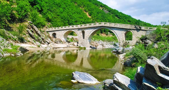 photo of Traditional stone arched bridge over river in Xanthi, Thrace, Greece. Vivid nature landscape, bridge reflecting on river water.
