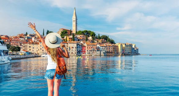 Photo of young tourist woman on harbour holding her hand up wave to beautiful Rovinj city, Croatia.