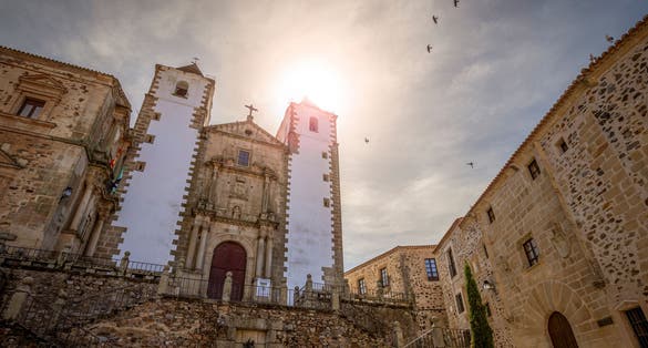 Photo of vView of the baroque church of San Francisco Javier de Caceres, Spain, in the Plaza de San Jorge at sunset with the sun backlit and pigeons in the sky .