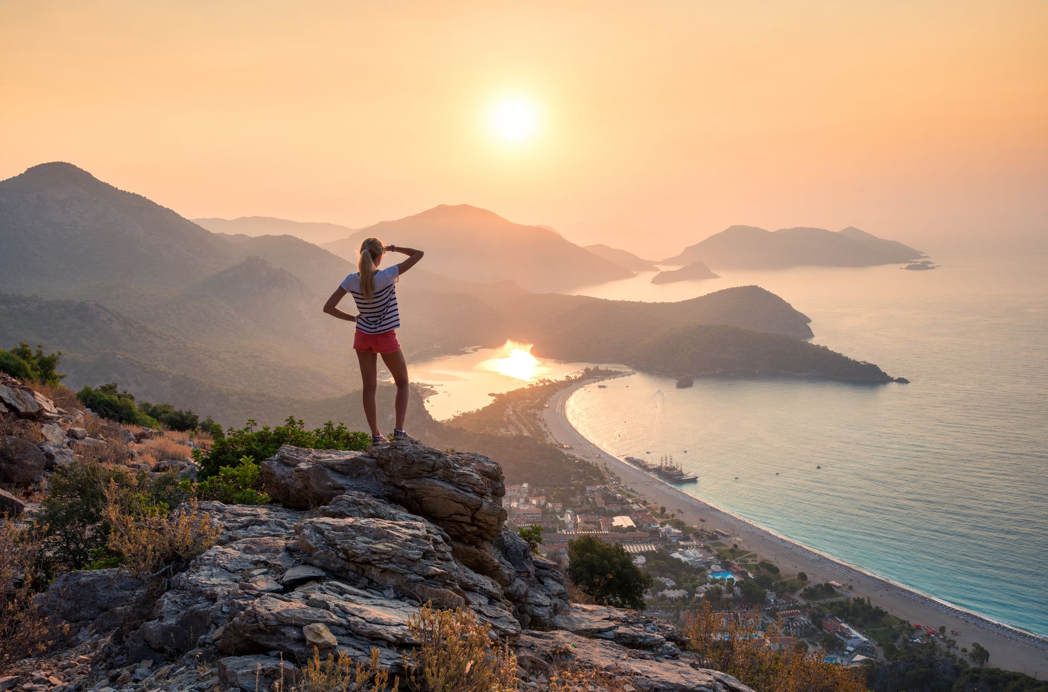 photo of young woman standing on the top of the rock and looking at the seashore and mountains at colorful sunset in summer in Ölüdeniz-Kıdrak Nature Park, Turkey.