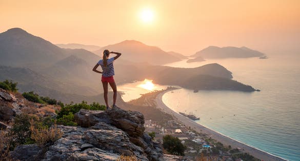 photo of young woman standing on the top of the rock and looking at the seashore and mountains at colorful sunset in summer in Ölüdeniz-Kıdrak Nature Park, Turkey.