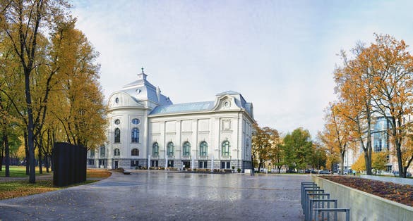Latvian National Museum of Art in Riga, a white historic building with arched windows and a metal roof, surrounded by autumn trees and an open paved square.
