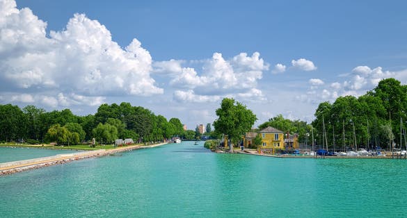 photo of view of View to entrance to Siofok harbor at Balaton lake, Hungary, with crystal clear water of emerald color, green coastline, puffy clouds on sky.