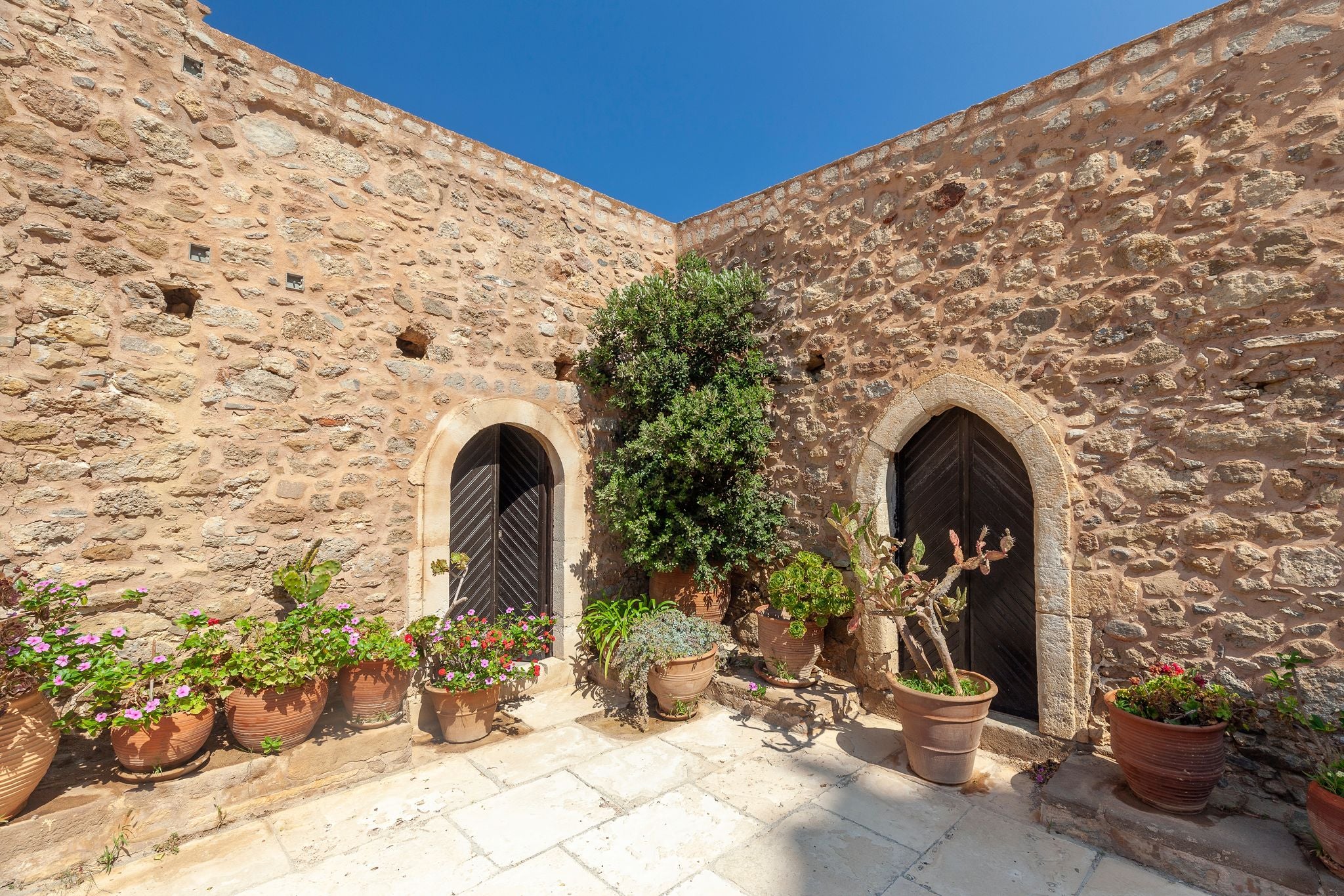 photo of view of Charming courtyard at the Moni Toplou Monastery in Sitia, Crete, adorned with vibrant potted plants against rustic stone walls under a clear blue sky.