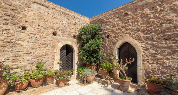 photo of view of Charming courtyard at the Moni Toplou Monastery in Sitia, Crete, adorned with vibrant potted plants against rustic stone walls under a clear blue sky.