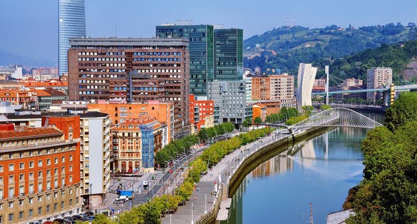 Photo of aerial view of Bilbao, Spain city downtown with a Nevion River, Zubizuri Bridge and promenade. Mountain at the background, with clear blue sky.