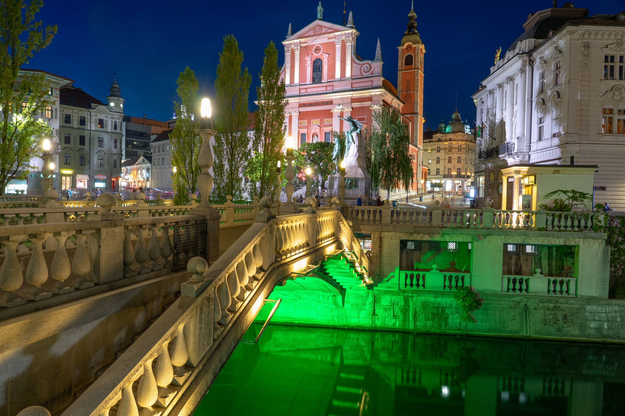 Prešeren Square with Tromostovje bridge on Ljubljanica river at night with city lights in Slovenia Ljubjana with Cerkev Marijinega oznanjenja church .