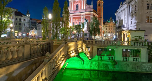 Prešeren Square with Tromostovje bridge on Ljubljanica river at night with city lights in Slovenia Ljubjana with Cerkev Marijinega oznanjenja church .